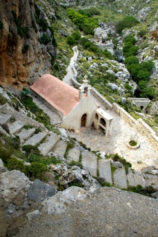 A small chapel with a red roof set in a rocky, mountainous landscape. Stone steps lead down to the chapel, surrounded by rich greenery and rugged terrain. A winding path continues into the distance, offering a sense of seclusion and tranquility.