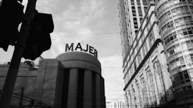 An urban scene depicting towering, classic-style buildings with a prominent sign that reads 'MAJES' on one of them. Power lines crisscross the view, adding to the metropolitan atmosphere. The sky is partly cloudy, casting a moody tone over the architectural structures.