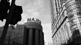 An urban scene depicting towering, classic-style buildings with a prominent sign that reads 'MAJES' on one of them. Power lines crisscross the view, adding to the metropolitan atmosphere. The sky is partly cloudy, casting a moody tone over the architectural structures.