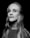 A black and white portrait of a person with long, straight hair and a neutral expression. The individual is wearing a shirt with visible branding on the shoulder. The lighting creates a subtle contrast, highlighting the contours of the face against a dark background.