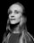 A black and white portrait of a person with long, straight hair and a neutral expression. The individual is wearing a shirt with visible branding on the shoulder. The lighting creates a subtle contrast, highlighting the contours of the face against a dark background.