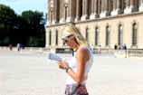 A woman with blonde hair wearing sunglasses and a white sleeveless top is reading a guidebook. She stands in front of a historical building with tall columns and arched windows. Other people are visible in the background, walking near the building.