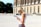 A woman with blonde hair wearing sunglasses and a white sleeveless top is reading a guidebook. She stands in front of a historical building with tall columns and arched windows. Other people are visible in the background, walking near the building.