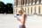 A woman with blonde hair wearing sunglasses and a white sleeveless top is reading a guidebook. She stands in front of a historical building with tall columns and arched windows. Other people are visible in the background, walking near the building.