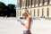 A woman with blonde hair wearing sunglasses and a white sleeveless top is reading a guidebook. She stands in front of a historical building with tall columns and arched windows. Other people are visible in the background, walking near the building.