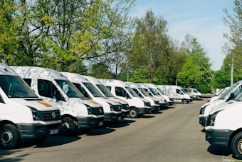 A row of uniformly parked white delivery vans on an asphalt surface with lush green trees in the background under a clear blue sky.