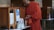 A person dressed in an orange robe is standing next to a wooden donation box with a transparent front, holding a plastic bag filled with cash. The setting appears to be indoors with intricately patterned walls and additional donation boxes in the background.