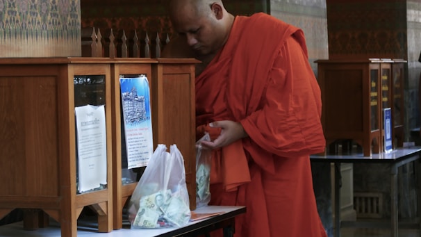 A person dressed in an orange robe is standing next to a wooden donation box with a transparent front, holding a plastic bag filled with cash. The setting appears to be indoors with intricately patterned walls and additional donation boxes in the background.