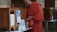 A person dressed in an orange robe is standing next to a wooden donation box with a transparent front, holding a plastic bag filled with cash. The setting appears to be indoors with intricately patterned walls and additional donation boxes in the background.