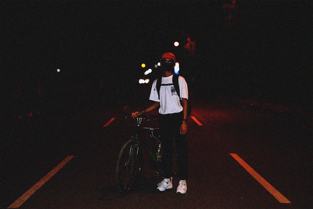 man in white t-shirt and black pants standing beside bicycle during nighttime