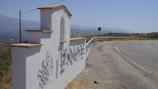 A white stucco welcome sign with the name 'TIZNATE' in large metallic letters casts a shadow on the ground. The sign has a stepped design with terracotta tiles on top, located beside a curving road with a distant view of mountains under a blue sky.