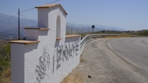 A white stucco welcome sign with the name 'TIZNATE' in large metallic letters casts a shadow on the ground. The sign has a stepped design with terracotta tiles on top, located beside a curving road with a distant view of mountains under a blue sky.