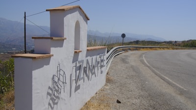 A white stucco welcome sign with the name 'TIZNATE' in large metallic letters casts a shadow on the ground. The sign has a stepped design with terracotta tiles on top, located beside a curving road with a distant view of mountains under a blue sky.