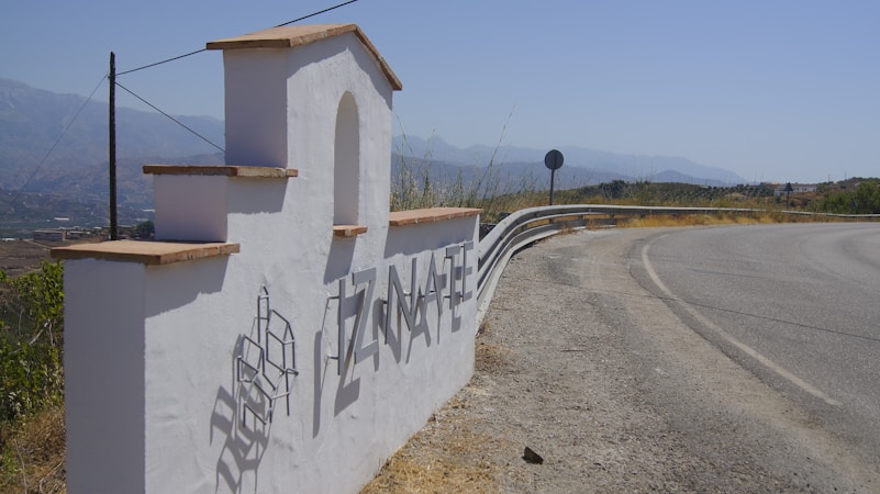 A white stucco welcome sign with the name 'TIZNATE' in large metallic letters casts a shadow on the ground. The sign has a stepped design with terracotta tiles on top, located beside a curving road with a distant view of mountains under a blue sky.