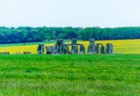 A historic site with large stone structures surrounded by lush green fields and a backdrop of dense trees. A group of people, possibly tourists, are gathered near the structures.