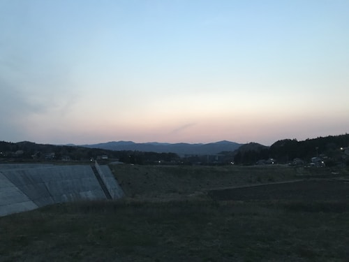 The image portrays a serene landscape at dusk or dawn, with a dimly lit sky transitioning through shades of blue and orange. Rolling hills line the horizon, and a flat, grassy field occupies the foreground. In the middle ground, there is a concrete embankment with a small set of stairs leading up to it.