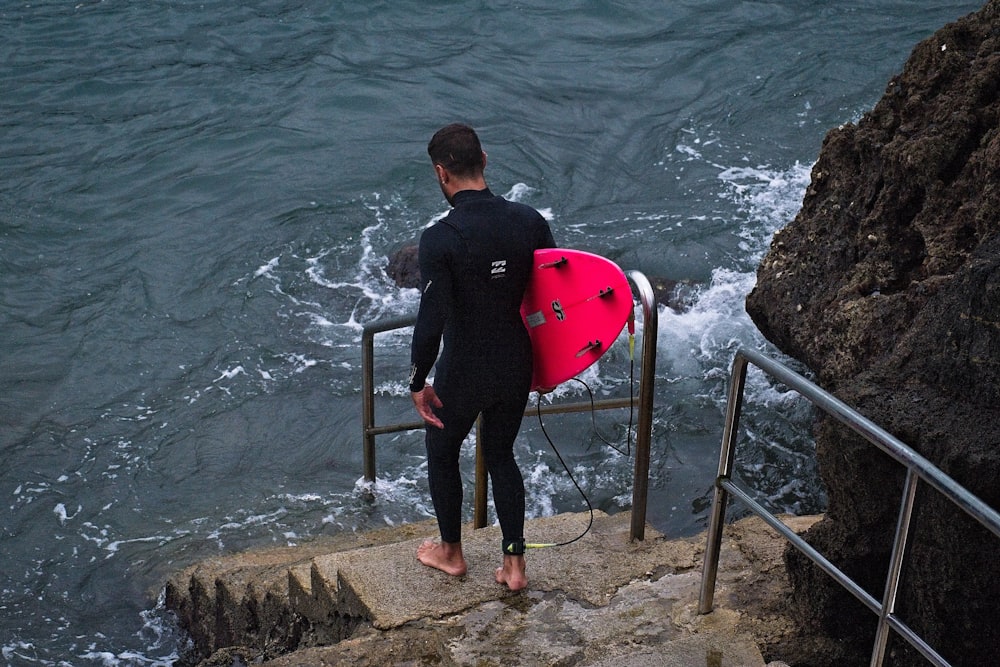 man in black jacket and pants holding pink and white surfboard standing on brown rock near near near near near