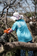 A person wearing a wide-brimmed hat and a blue t-shirt with a logo engages in a cleanup effort in a wooded area. They are pulling on some debris with orange gloves, surrounded by leafless trees.