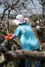 A person wearing a wide-brimmed hat and a blue t-shirt with a logo engages in a cleanup effort in a wooded area. They are pulling on some debris with orange gloves, surrounded by leafless trees.