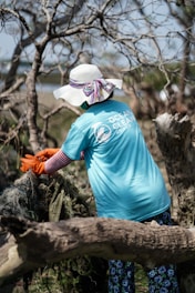 A person wearing a wide-brimmed hat and a blue t-shirt with a logo engages in a cleanup effort in a wooded area. They are pulling on some debris with orange gloves, surrounded by leafless trees.