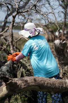 A person wearing a wide-brimmed hat and a blue t-shirt with a logo engages in a cleanup effort in a wooded area. They are pulling on some debris with orange gloves, surrounded by leafless trees.
