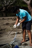 A person wearing a blue shirt and gloves is cleaning up a beach area, handling a stick and a pile of tangled debris, including nets or ropes. The background features trees and sparse vegetation.