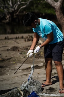 A person wearing a blue shirt and gloves is cleaning up a beach area, handling a stick and a pile of tangled debris, including nets or ropes. The background features trees and sparse vegetation.