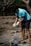 A person wearing a blue shirt and gloves is cleaning up a beach area, handling a stick and a pile of tangled debris, including nets or ropes. The background features trees and sparse vegetation.