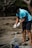 A person wearing a blue shirt and gloves is cleaning up a beach area, handling a stick and a pile of tangled debris, including nets or ropes. The background features trees and sparse vegetation.