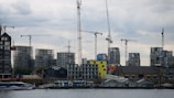 A waterfront construction site features several tower cranes and partially built high-rise buildings, with a cloudy sky above and a modern glass building at the water's edge.