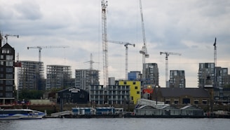 A waterfront construction site features several tower cranes and partially built high-rise buildings, with a cloudy sky above and a modern glass building at the water's edge.