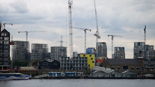 A waterfront construction site features several tower cranes and partially built high-rise buildings, with a cloudy sky above and a modern glass building at the water's edge.