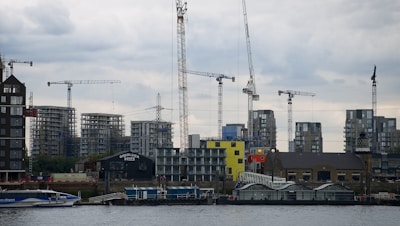A waterfront construction site features several tower cranes and partially built high-rise buildings, with a cloudy sky above and a modern glass building at the water's edge.