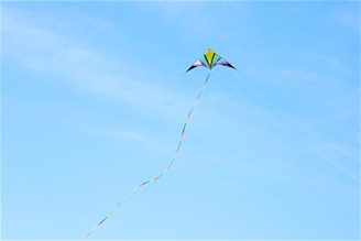 A colorful kite with a long, multicolored tail soars against a clear blue sky. The kite has a symmetrical triangular shape with vibrant colors including yellow, purple, and green.