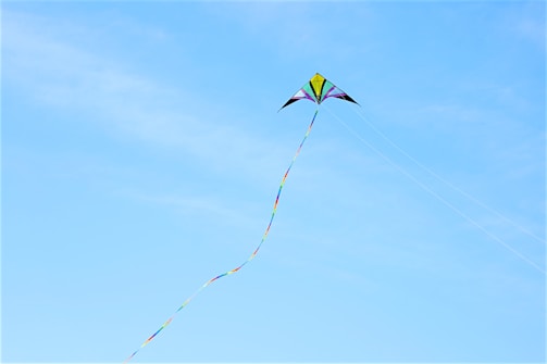 A colorful kite with a long, multicolored tail soars against a clear blue sky. The kite has a symmetrical triangular shape with vibrant colors including yellow, purple, and green.
