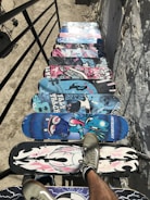A series of colorful skateboard decks are lined up as steps on a staircase. The decks feature a variety of vibrant designs, including skulls, cartoon characters, and abstract patterns. There are two legs with sneakers visible on one of the decks, giving the impression of someone walking down the stairway of skateboards. The setting is outdoor, and the surrounding surfaces are rough and textured.