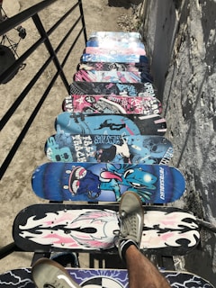 A series of colorful skateboard decks are lined up as steps on a staircase. The decks feature a variety of vibrant designs, including skulls, cartoon characters, and abstract patterns. There are two legs with sneakers visible on one of the decks, giving the impression of someone walking down the stairway of skateboards. The setting is outdoor, and the surrounding surfaces are rough and textured.