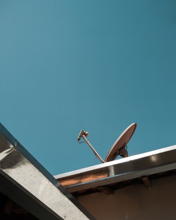 A satellite dish is mounted on a rooftop under a clear blue sky. The structure of the roof and the mounting hardware are visible, with a minimalistic and uncluttered appearance.