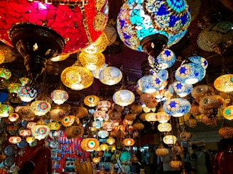 A vibrant display of multicolored, intricately designed mosaic lamps hanging in a market setting. The lamps are of various shapes and sizes, showcasing patterns in bright reds, blues, yellows, and greens. The warm glow from the lamps creates a lively and enchanting atmosphere.
