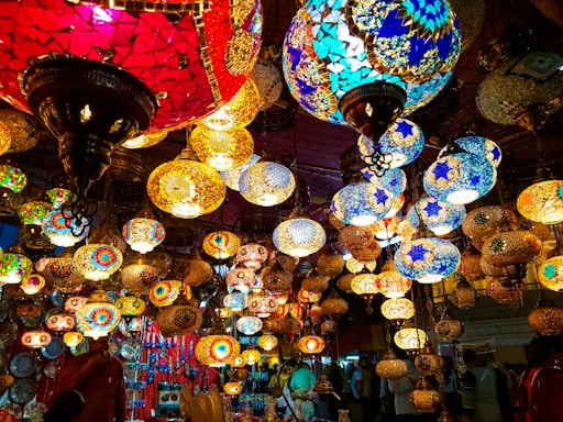 A vibrant display of multicolored, intricately designed mosaic lamps hanging in a market setting. The lamps are of various shapes and sizes, showcasing patterns in bright reds, blues, yellows, and greens. The warm glow from the lamps creates a lively and enchanting atmosphere.