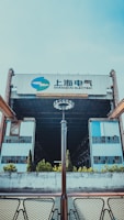 A large industrial building with the words 'Shanghai Electric' displayed prominently atop. The structure features metal and glass elements, creating a sense of industrialism. The foreground includes a symmetrical arrangement of modern light fixtures and patio furniture, with small bushes and trees lining the front approach.