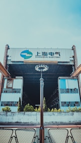 A large industrial building with the words 'Shanghai Electric' displayed prominently atop. The structure features metal and glass elements, creating a sense of industrialism. The foreground includes a symmetrical arrangement of modern light fixtures and patio furniture, with small bushes and trees lining the front approach.