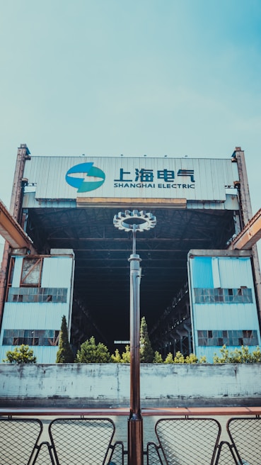 A large industrial building with the words 'Shanghai Electric' displayed prominently atop. The structure features metal and glass elements, creating a sense of industrialism. The foreground includes a symmetrical arrangement of modern light fixtures and patio furniture, with small bushes and trees lining the front approach.