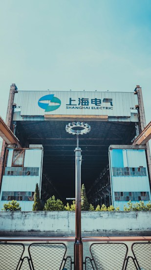 A large industrial building with the words 'Shanghai Electric' displayed prominently atop. The structure features metal and glass elements, creating a sense of industrialism. The foreground includes a symmetrical arrangement of modern light fixtures and patio furniture, with small bushes and trees lining the front approach.