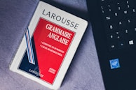 A paperback book titled 'Grammaire Anglaise' by Larousse lies on a soft, gray surface next to a black laptop keyboard with visible Intel logo. The book has a red, white, and blue cover with text in French emphasizing essential grammar modules.