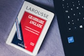 A paperback book titled 'Grammaire Anglaise' by Larousse lies on a soft, gray surface next to a black laptop keyboard with visible Intel logo. The book has a red, white, and blue cover with text in French emphasizing essential grammar modules.