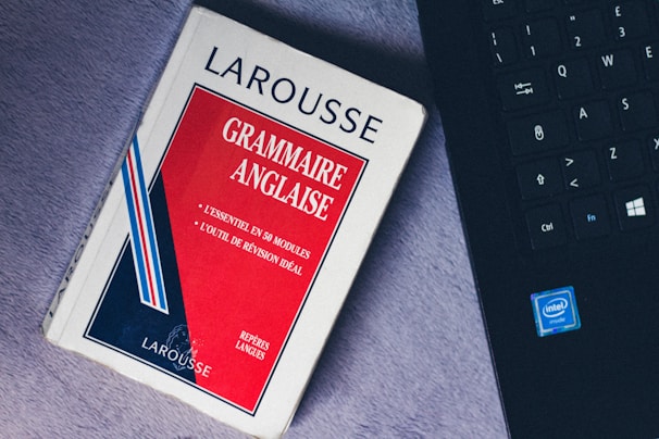 A paperback book titled 'Grammaire Anglaise' by Larousse lies on a soft, gray surface next to a black laptop keyboard with visible Intel logo. The book has a red, white, and blue cover with text in French emphasizing essential grammar modules.