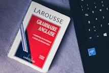 A paperback book titled 'Grammaire Anglaise' by Larousse lies on a soft, gray surface next to a black laptop keyboard with visible Intel logo. The book has a red, white, and blue cover with text in French emphasizing essential grammar modules.