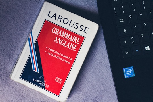 A paperback book titled 'Grammaire Anglaise' by Larousse lies on a soft, gray surface next to a black laptop keyboard with visible Intel logo. The book has a red, white, and blue cover with text in French emphasizing essential grammar modules.