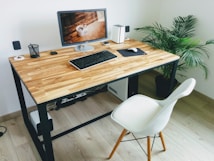 A modern workspace featuring a wooden desk with a computer monitor, keyboard, and mouse. On the desk are small speakers, a pen holder, and a decorative plant to the side. The room is minimalistic with a white chair and a potted plant in the corner.